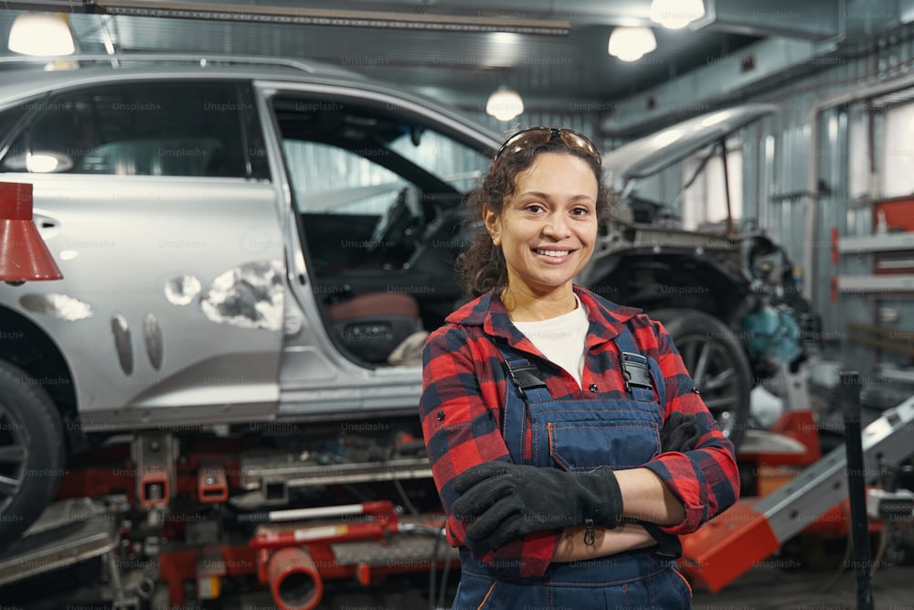 Fröhliche Arbeiterin, die die Arme verschränkt hält und lächelt, während sie in der Nähe des Fahrzeugs in der Reparaturwerkstatt steht