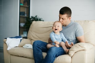 Affectionate father kissing his happy baby boy while relaxing on the sofa at home.