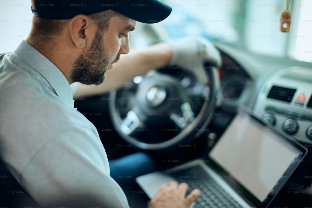 Trabalhador do sexo masculino usando laptop enquanto faz diagnóstico de carro e trabalha em oficina de reparação de automóveis.
