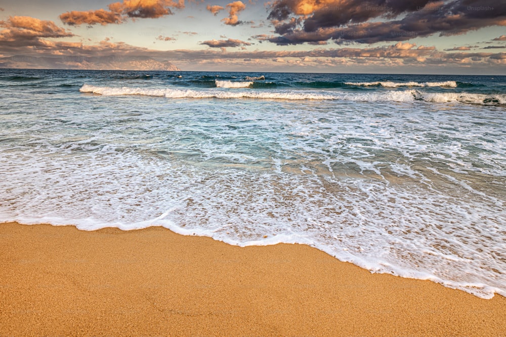 Weitwinkelansicht eines idyllischen Meeressandstrandes mit einer ankommenden azurblauen Welle bei Flut. Windiges und stürmisches Wetter bei Sonnenuntergang. Hintergrund für Reisen und Urlaub
