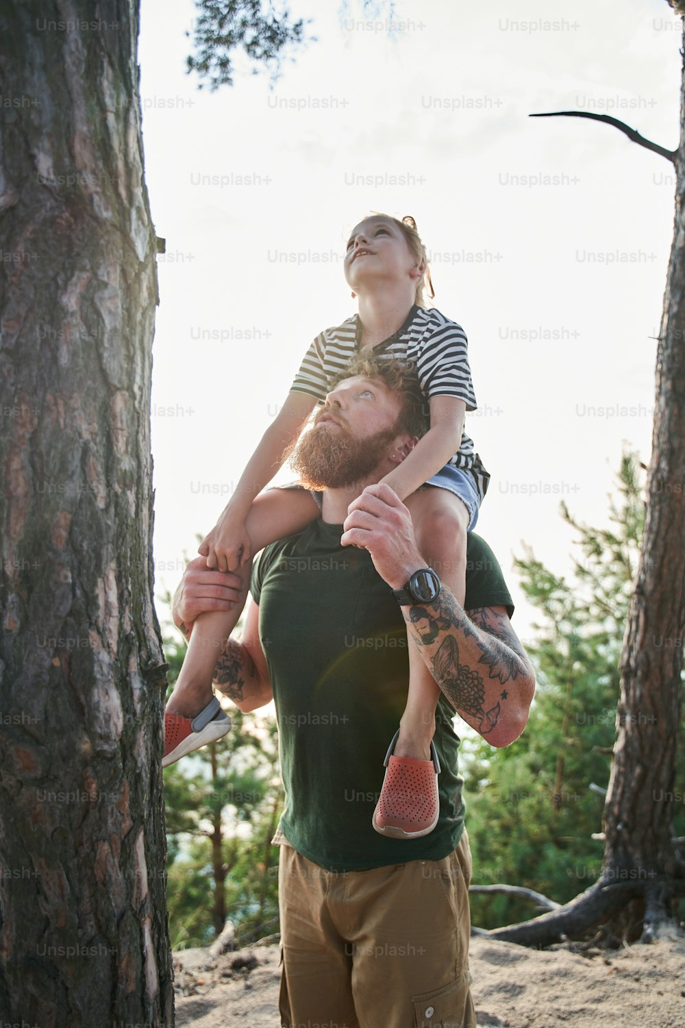 Attractive young man is playing with his daughter in the nature. Father is standing and carrying girl on his back. They looking at the tree