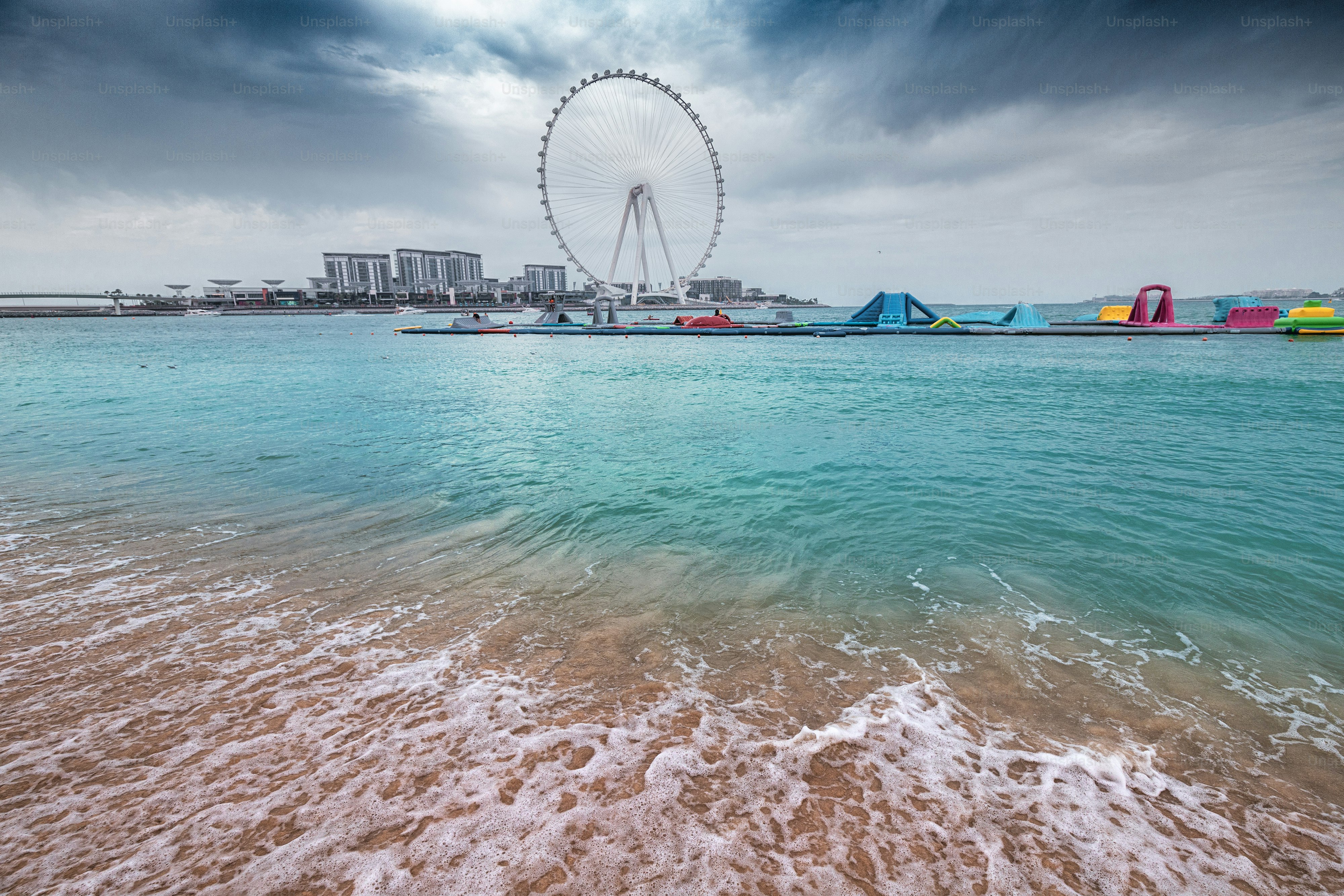 A wave crashes on the sandy beach and the famous Dubai Eye Ferris Wheel during cloudy weather