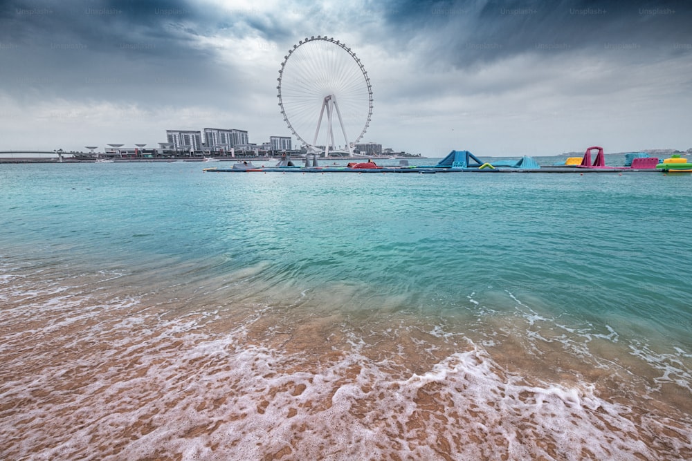 A wave crashes on the sandy beach and the famous Dubai Eye Ferris Wheel during cloudy weather