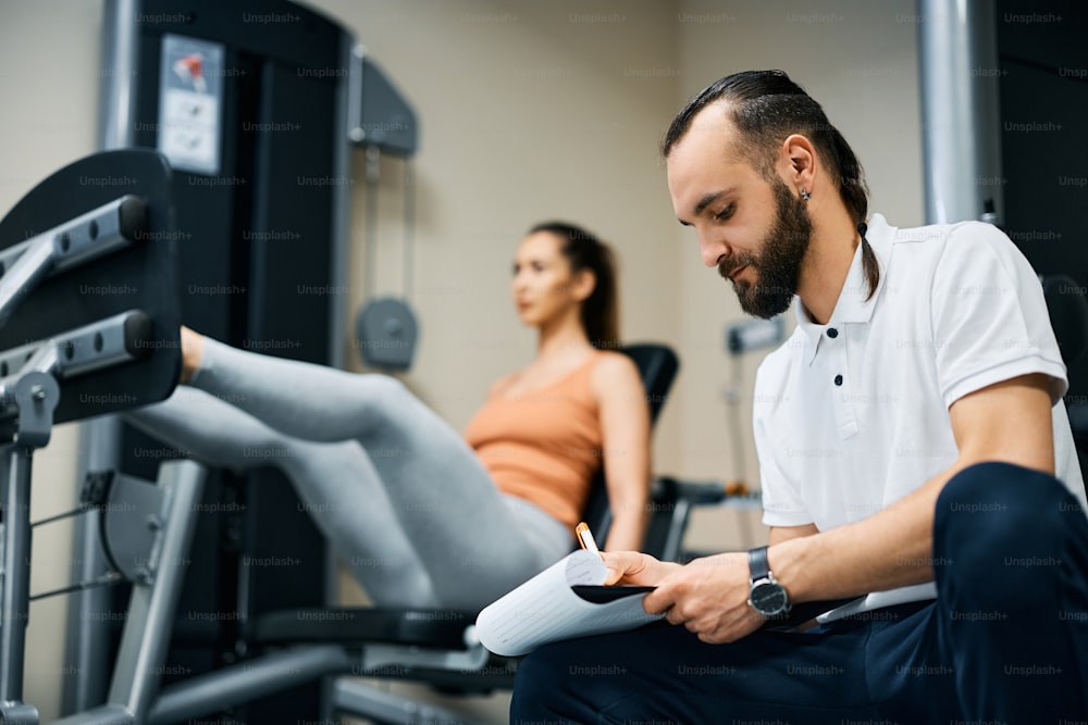 Entrenador personal escribiendo en el portapapeles mientras la deportista practica en la máquina de prensa de piernas en el gimnasio.