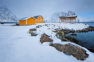 Traditionelles gelbes Rorbu-Haus in trocknenden Flocken für Stockfisch-Kabeljau im norwegischen Fjord im Winter. Fischerdorf Sakrisoy, Lofoten, Norwegen