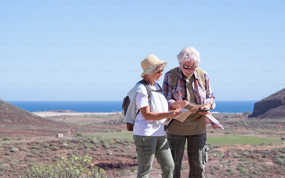 Active senior couple with backpacks in outdoors excursion hiking in mountain looking at map enjoying healthy lifestyle.  Scenic view of sea and mountain background