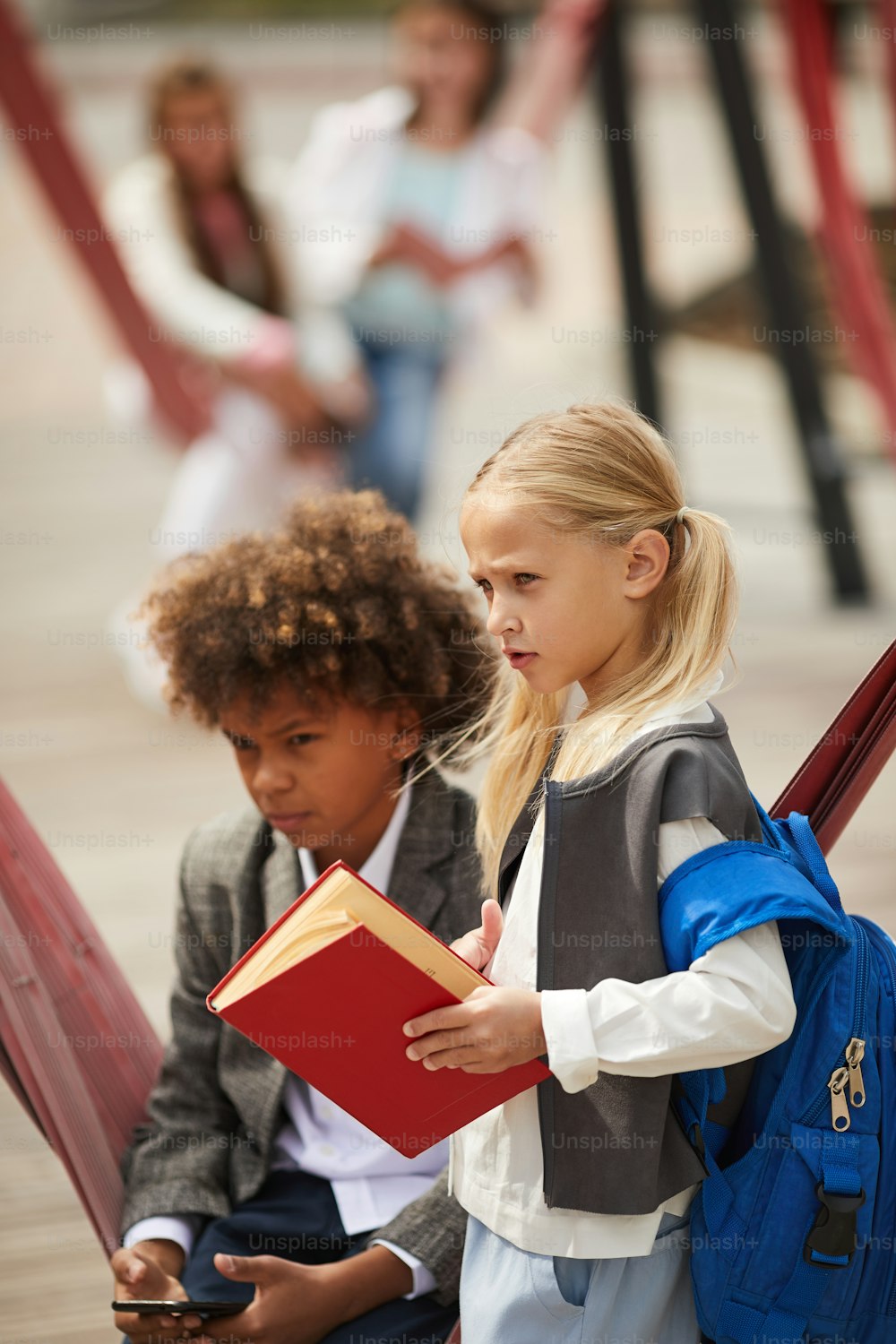 Blond schoolgirl with backpack behind her back holding book standing on the playground with African boy who using mobile phone