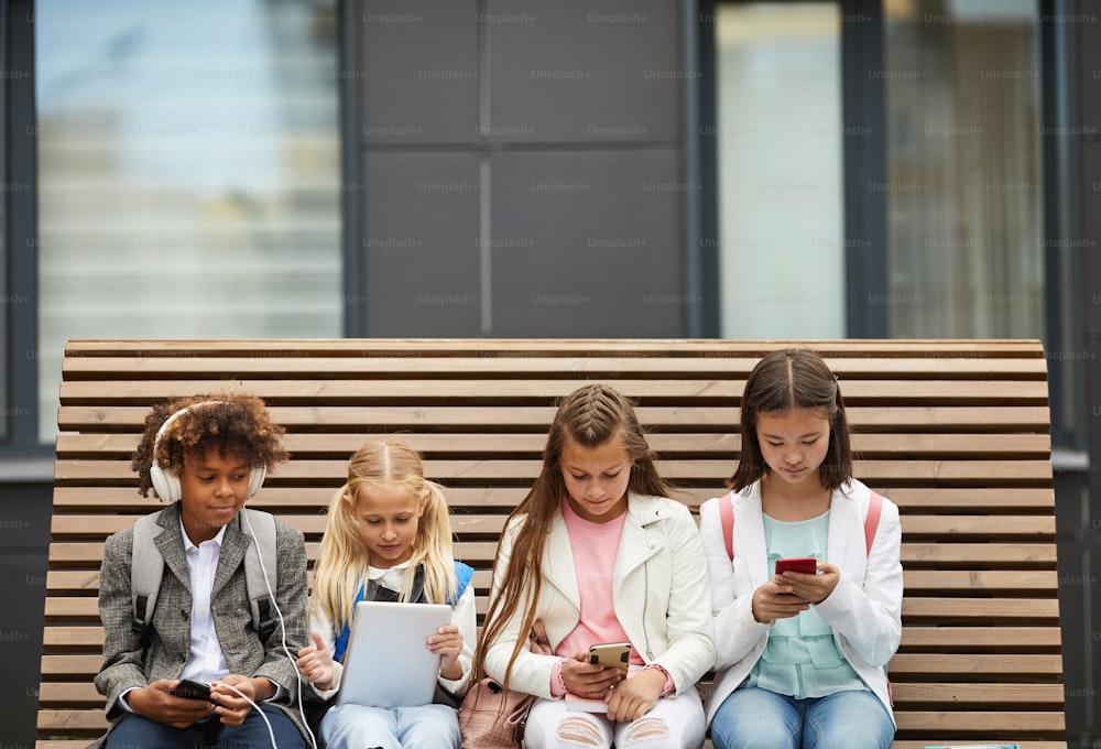Group of school children sitting on the bench and using different gadgets they studying online