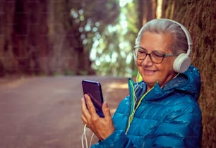 イヤホンをした魅力的な孤独な年配の女性が携帯電話のメッセージや音楽を聴いています。秋冬の季節に、自然の中でアウトドア
