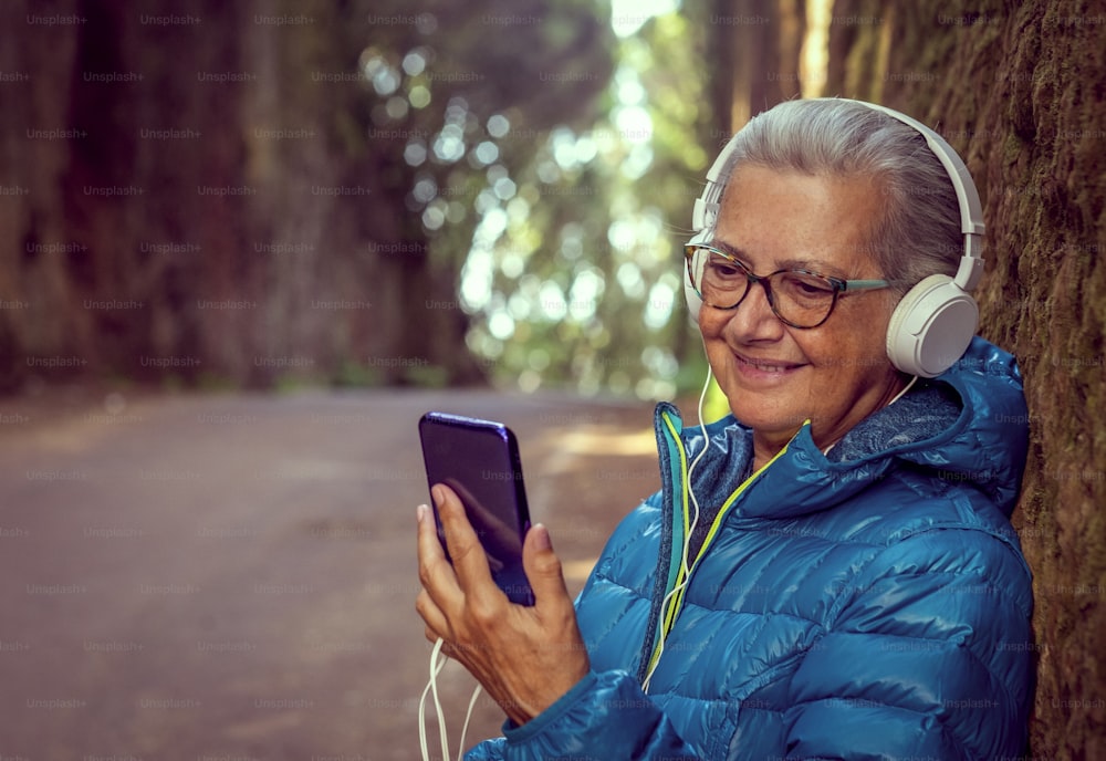 Attractive lonely elderly woman with earphones listens to mobile phone message or music. Outdoors in nature, in the autumn winter season