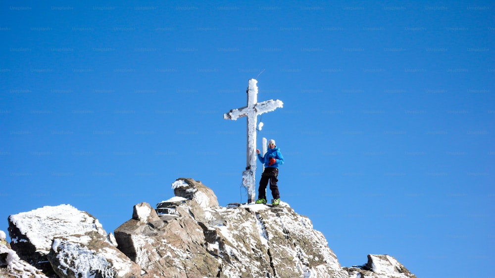 Ein männlicher Backcountry-Skifahrer am Gipfelkreuz eines hochalpinen Gipfels an einem schönen Wintertag