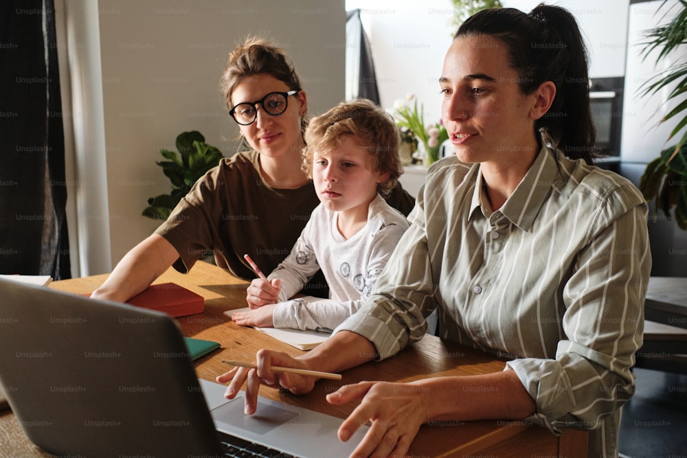 Madres lesbianas jóvenes usando una computadora portátil mientras están sentadas a la mesa con su hijo y lo ayudan con el estudio