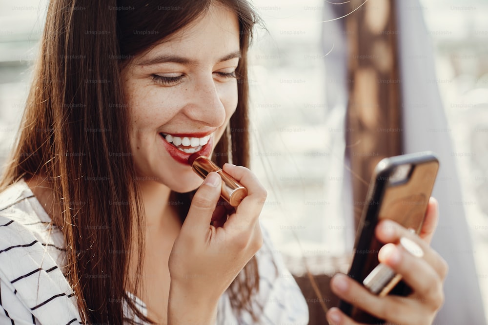 Beautiful stylish young woman applying red lipstick on lips and looking at phone screen. Happy hipster girl in retro dress relaxing and smiling on roof in sunny  european city