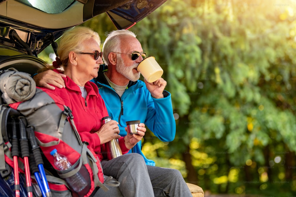 Senior couple sitting  in car trunk, drink coffee while hiking in the forest.