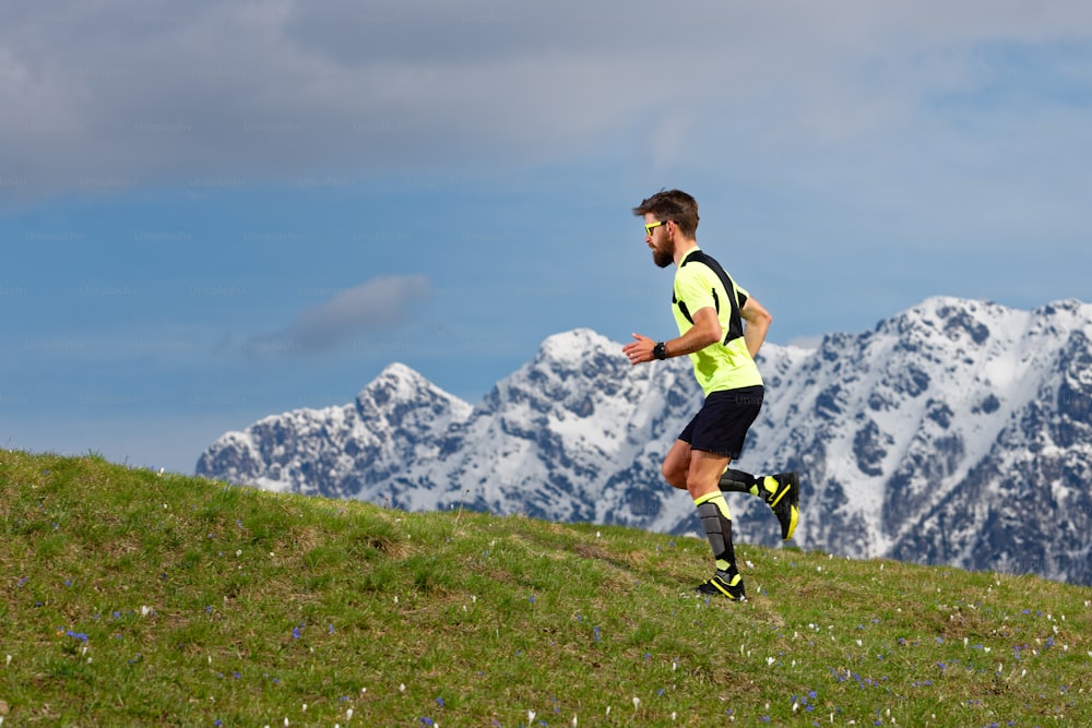 Skyrunning a bearded man in a spring meadow with snow mountains background.