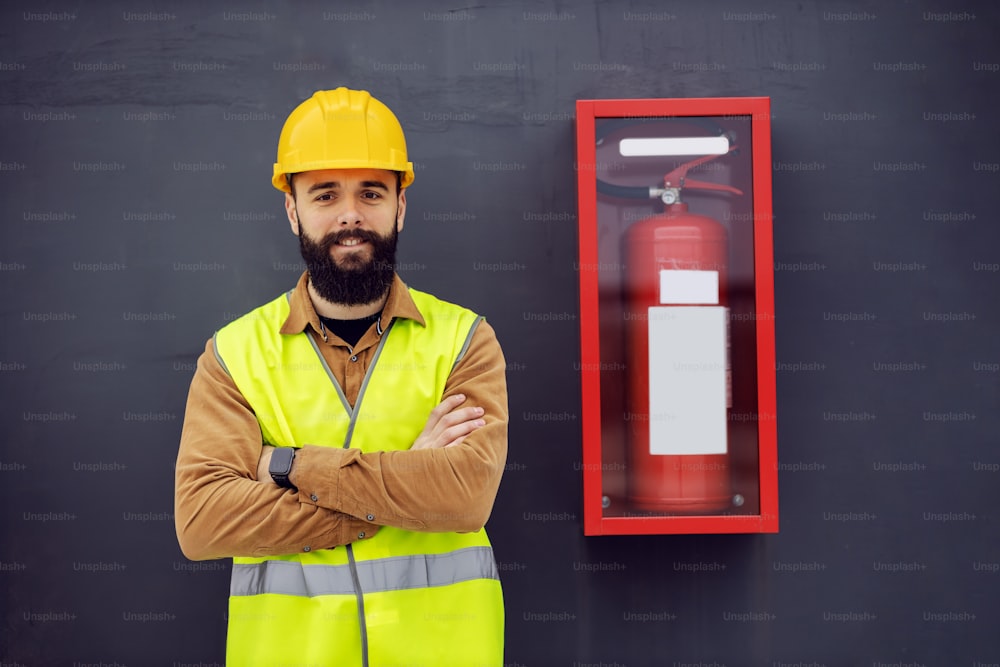 Young smiling cheerful attractive bearded worker in vest, with helmet on head standing next to fire fire extinguisher with arms crossed. Safety comes first.