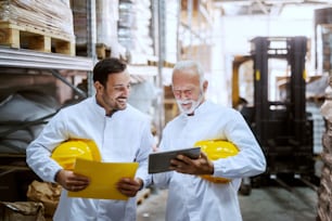 Smiling employees in white uniforms and yellow helmets under armpits standing in warehouse and comparing data from tablet. Senior worker holding tablet while younger one holding yellow folder.
