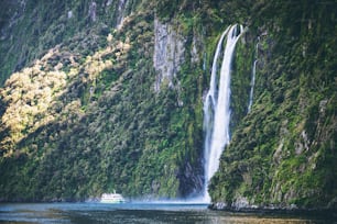 Sightseeing boat carrying tourist people approaches great waterfall in Milford Sound. Beautiful scenic cruise through Fiordland National Park in South Island of New Zealand.