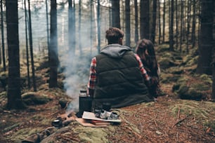 Rückansicht eines jungen Paares, das sich am Lagerfeuer aufwärmt, während es im Wald sitzt