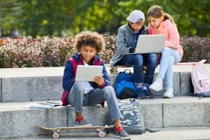 African boy using tablet pc while sitting on stairs with skateboard outdoors with his friends using laptop in the background