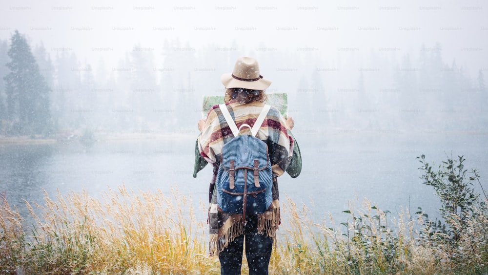 Travel woman reading map among amazing snowy wild, enjoy the nature landsape. Forest and lake, wearing hat and poncho, boho and wanderlust style