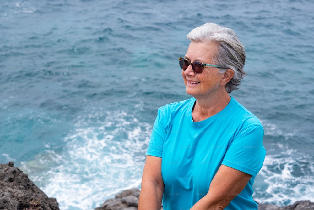 Senior woman in outdoor excursion at sea sitting on the cliff looking at horizon over water. Retirement lifestyle