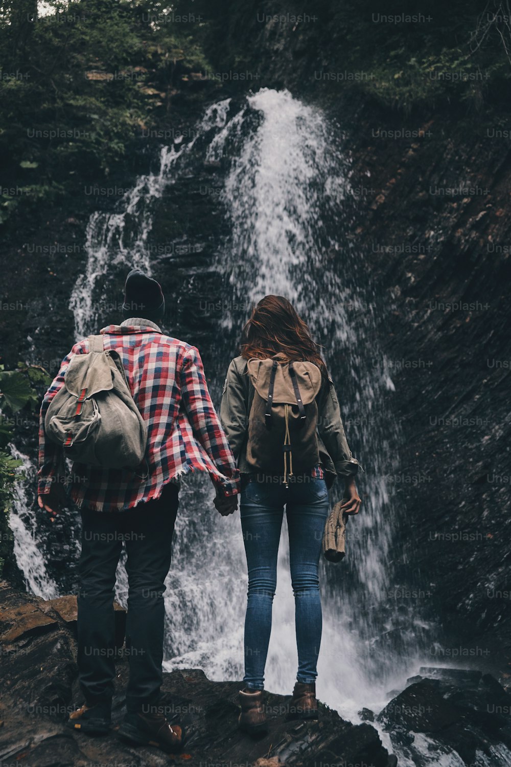 滝の近くに立っている間、手をつないでいる若いカップルの全身背面図