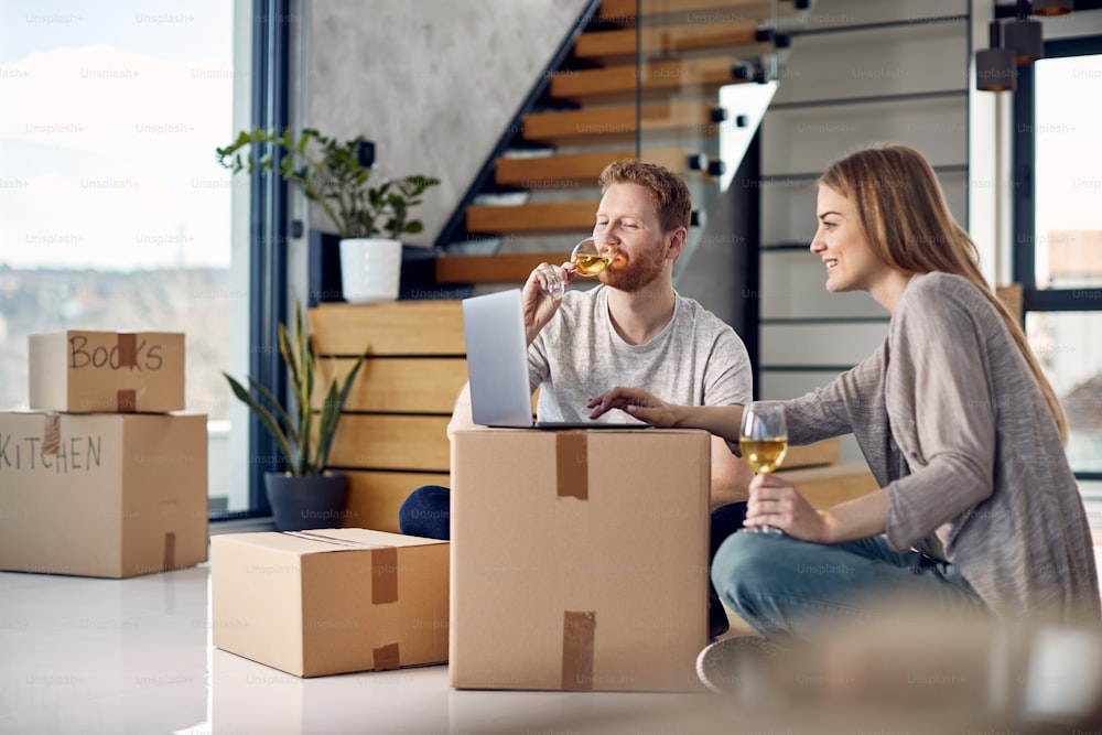 Couple souriant surfant sur le net sur un ordinateur portable et buvant un verre de vin tout en se relaxant sur le sol de leur nouvel appartement.