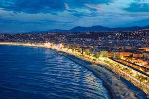 Vista panorâmica de Nice, França à noite hora azul. Ondas do Mar Mediterrâneo surgindo na costa, pessoas relaxando na praia, iluminação de luzes em casas coloridas