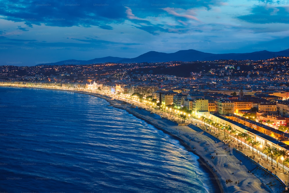 Vista panorâmica de Nice, França à noite hora azul. Ondas do Mar Mediterrâneo surgindo na costa, pessoas relaxando na praia, iluminação de luzes em casas coloridas