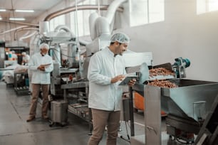 Jeune superviseur caucasien évaluant la qualité de la nourriture dans l’usine alimentaire tout en tenant une tablette. L’homme est vêtu d’un uniforme blanc et porte un filet à cheveux.