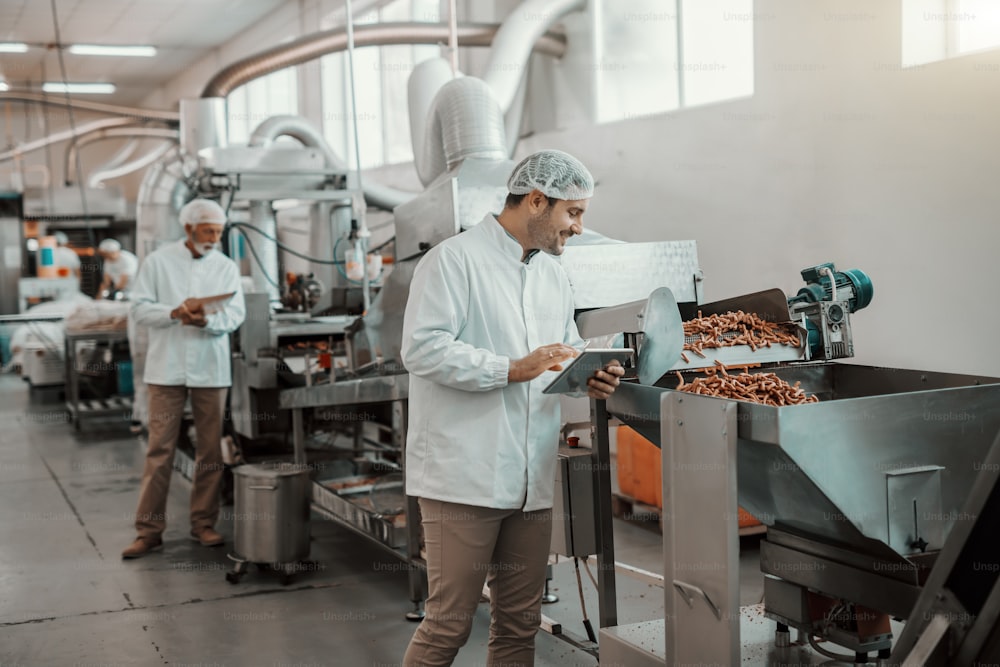 Young Caucasian supervisor evaluating quality of food in food plant while holding tablet. Man is dressed in white uniform and having hair net.