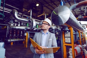 Guapo hombre de negocios caucásico de traje y con casco protector en la cabeza sosteniendo una carpeta con documentos mientras está de pie en la planta de energía.