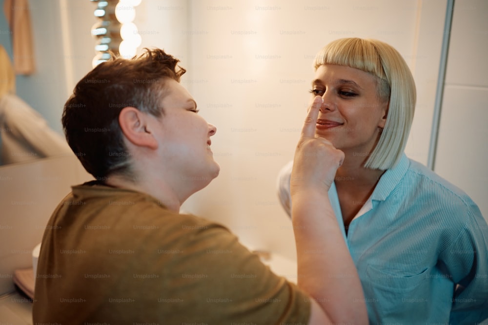 Junges lesbisches Paar hat Spaß bei der Verwendung von Gesichtscreme im Badezimmer.