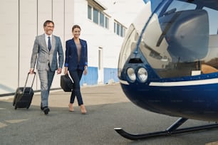 Cheerful businessman with trolley suitcase and smiling businesswoman with bag approaching chopper