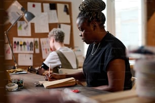 Ernsthafte afroamerikanische Schneiderin oder Designerin, die durch Lookbooks schaut und über neue Ideen für die Modekollektion nachdenkt