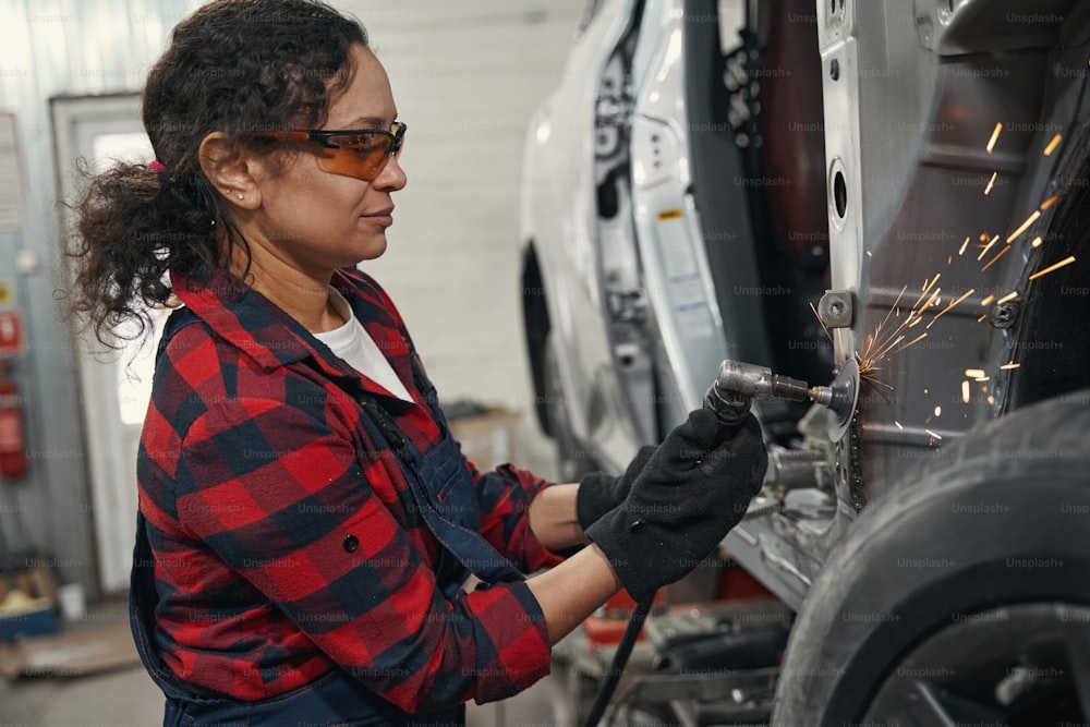Woman vehicle technician in safety glasses using grinding device while fixing car in auto repair service station