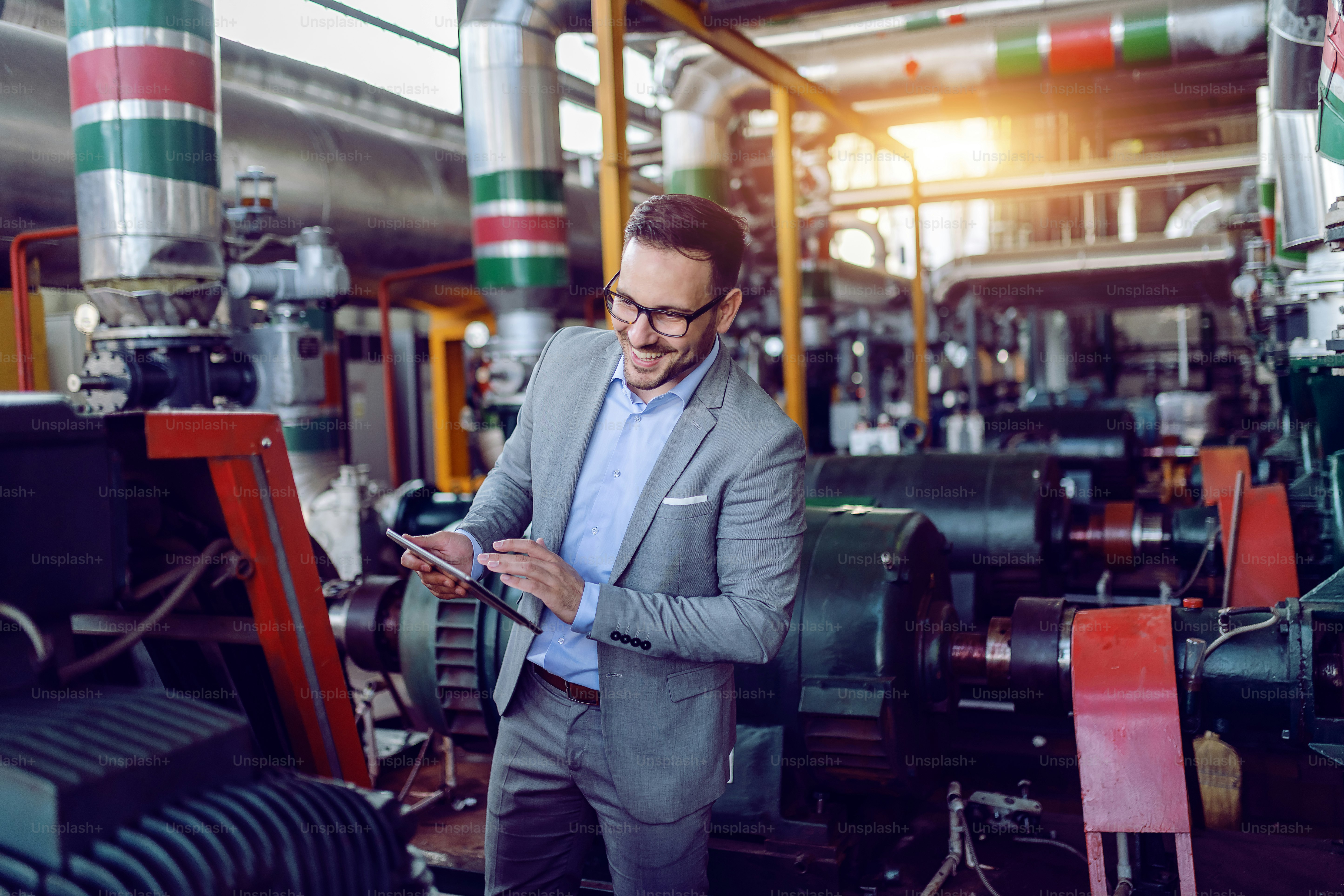 Smiling caucasian supervisor in suit holding tablet and checking on machinery in energy plant.