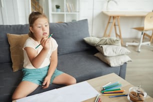 Girl with down syndrome sitting on sofa in front of the table she learning to draw with pencils at home