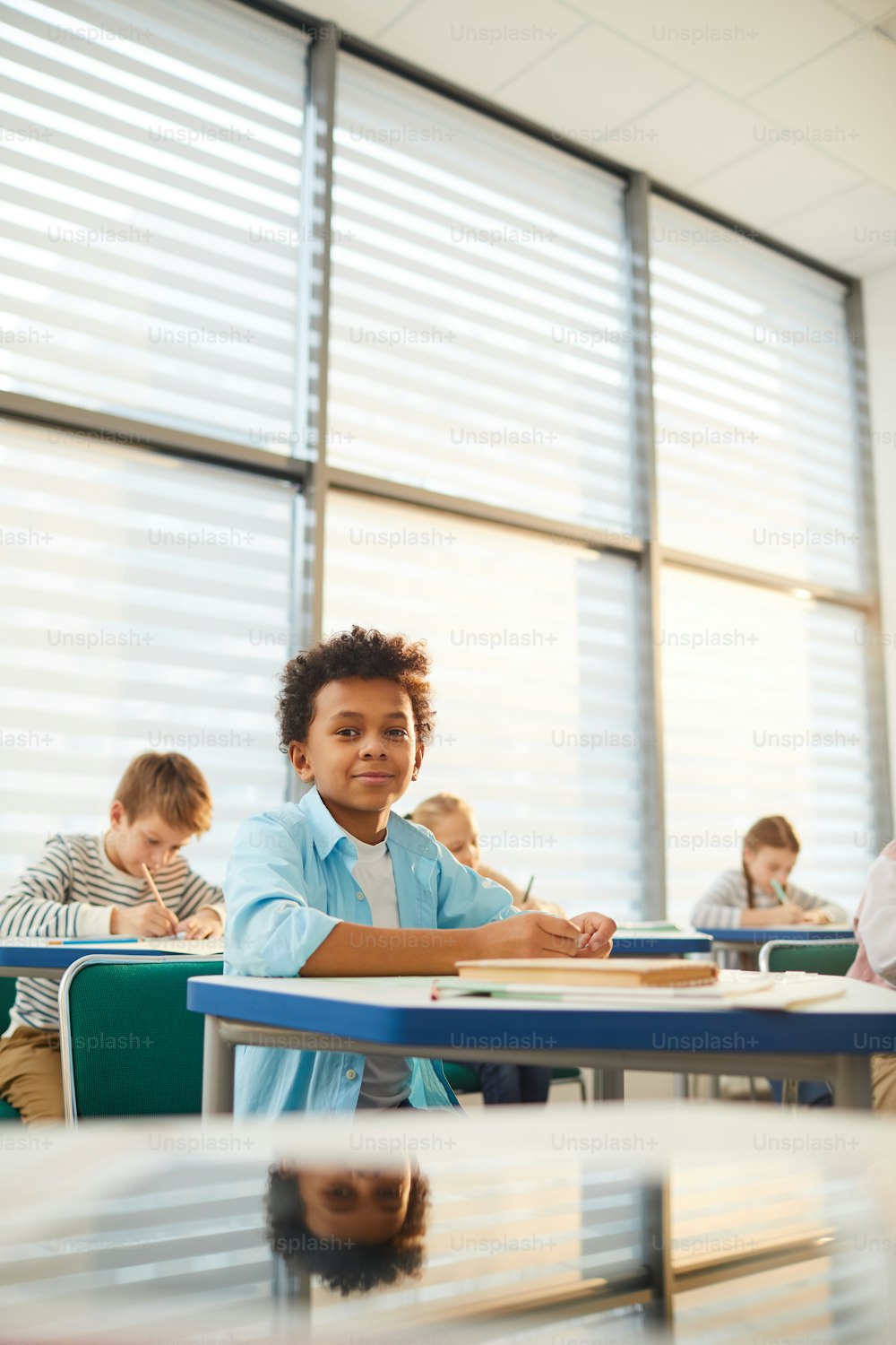 Foto vertical de estudante afro-americano sentado na mesa na sala de aula moderna olhando para a câmera enquanto faz o exercício da aula, espaço de cópia