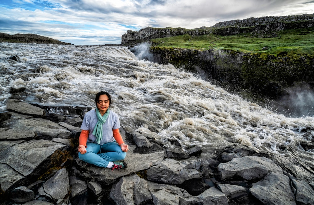 Young woman doing meditation yoga while sitting on side of river and large waterfall landscape of Dettifoss waterfall in Iceland.