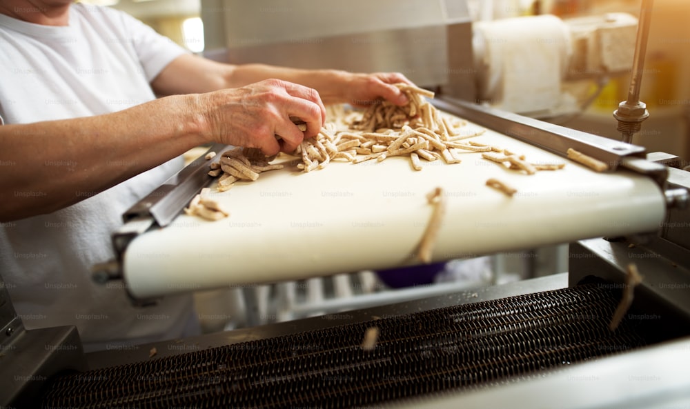 Mature careful dedicated worker is spreading salt stick dough across the healthy food production line conveyor for further processing.