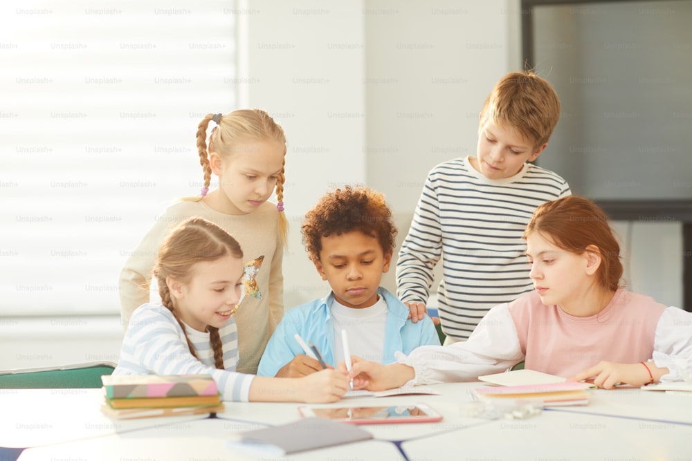 Duas meninas e meninos sentados juntos na sala de aula desenhando algo em um caderno, suas frends em pé atrás deles, tiro horizontal