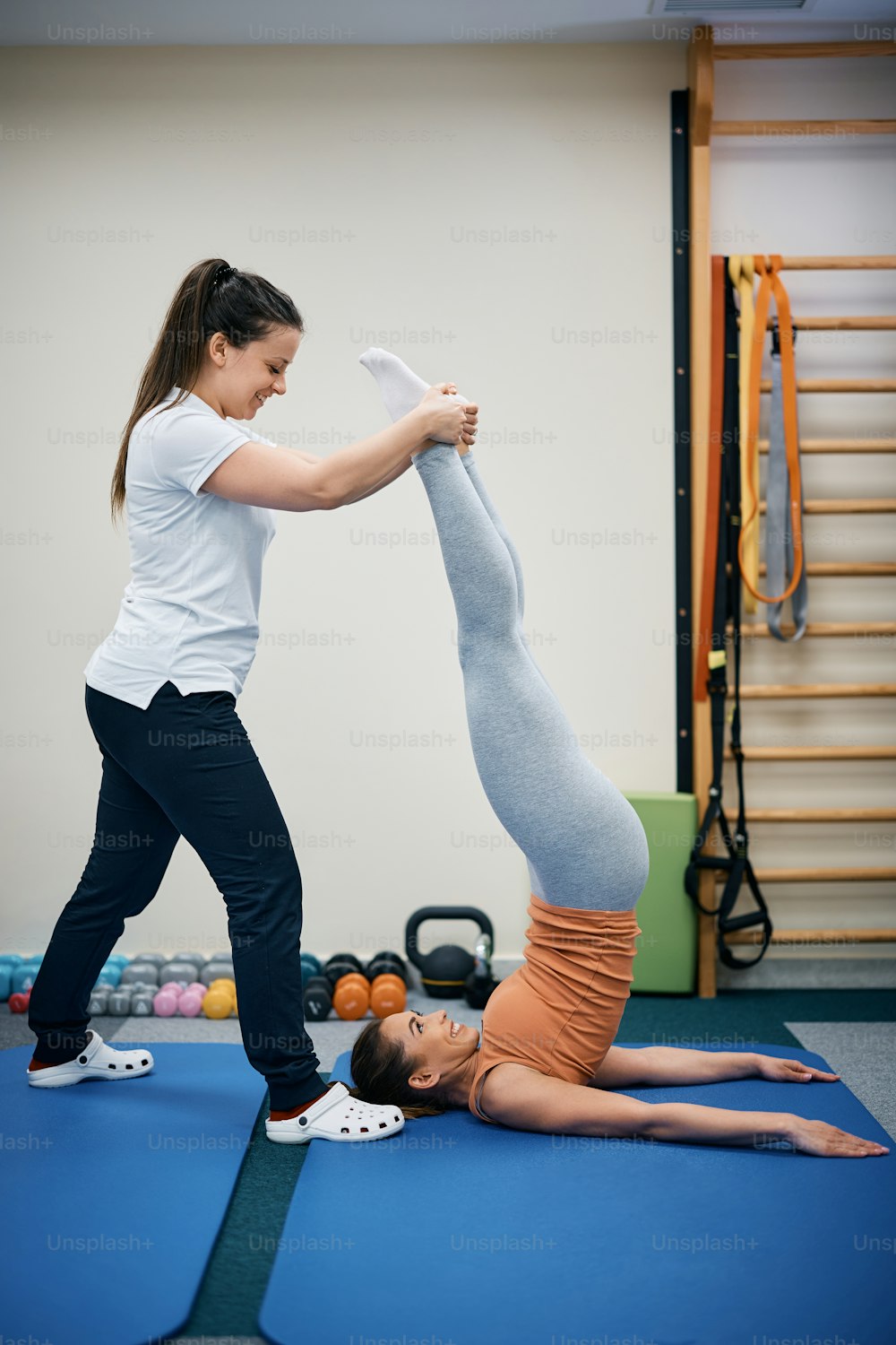 Happy physiotherapist assisting young woman with shoulder stand exercise during physical therapy treatment at health club.