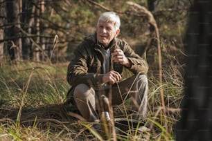 Full length view of the old male tourist looking at the nature while drinking beverage and warming after collecting mushrooms