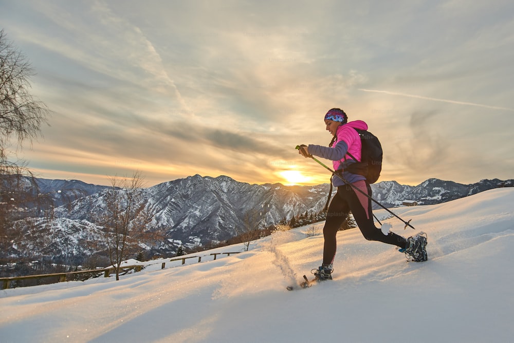 Mulher esportiva nova na neve com raquetes de neve em uma paisagem do pôr do sol