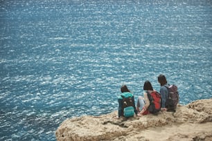 Paz e relaxamento. Mulheres jovens serenas estão sentadas na montanha rochosa e olhando para o mar.  Mochilas turísticas estão nas costas