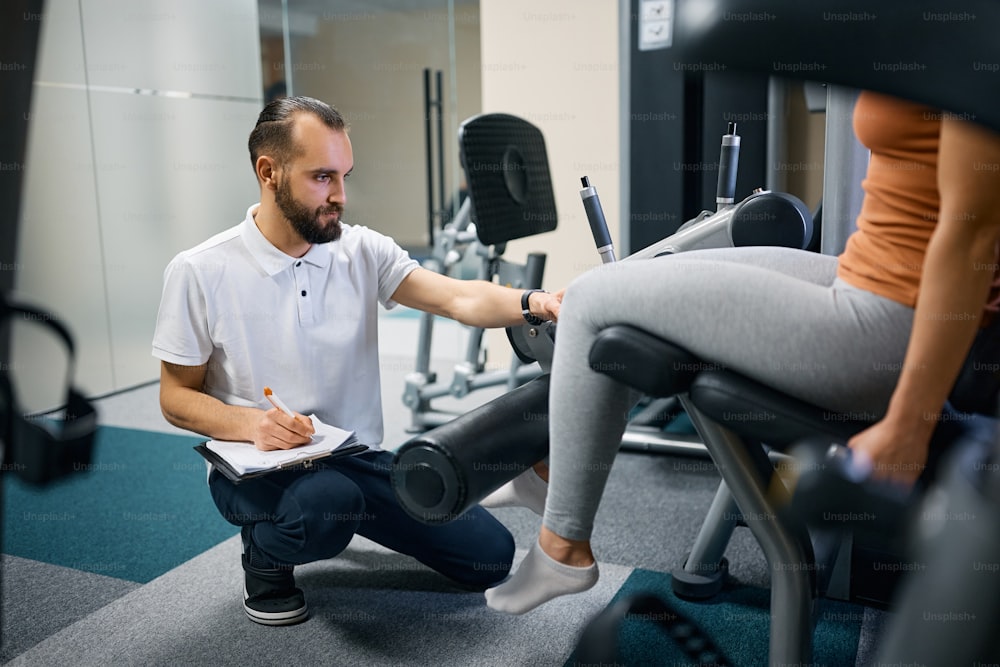 Physical therapist monitoring athletic woman who is practicing on leg extension machine during her rehabilitation treatment.