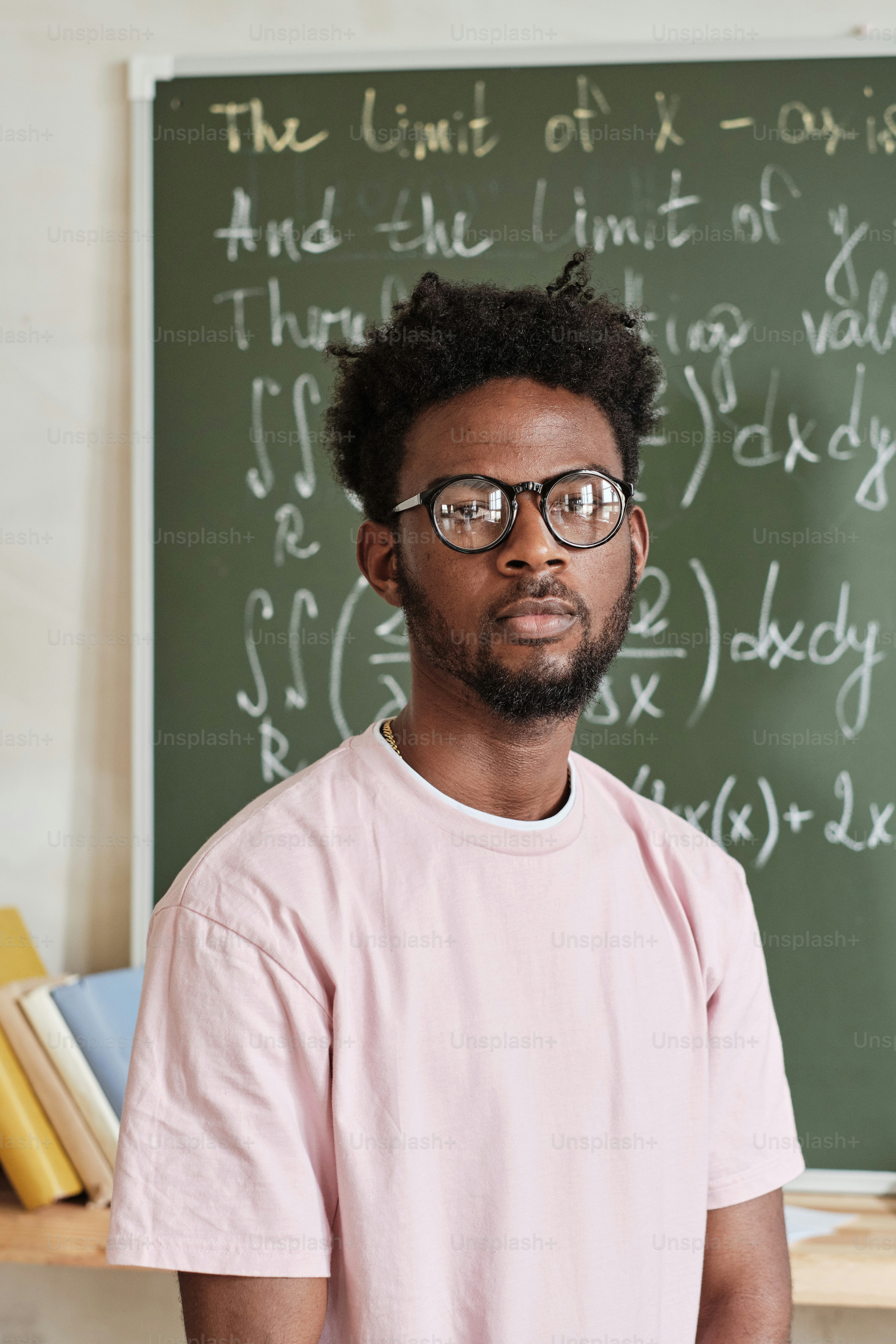 Portrait of African student in eyeglasses looking at camera while standing near the blackboard in the classroom