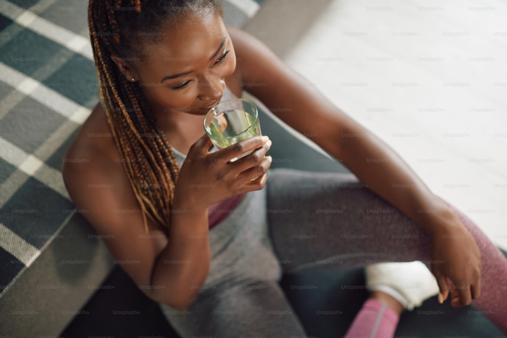 Young black sportswoman drinking water while taking a break from exercising at home.